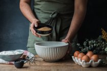 Unrecognizable man in apron spilling cane sugar into bowl near flour and eggs while preparing batter for pastry — Stock Photo