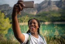 Encantada viajera afroamericana tomando uno mismo disparo en el teléfono inteligente en el fondo del lago en las tierras altas en verano - foto de stock