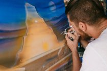 Вид збоку художника-чоловіка, який використовує пульверизатор для малювання картини на полотні під час роботи в творчій майстерні — стокове фото