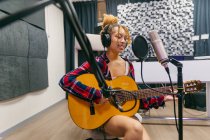Молода етнічна жінка-вокалістка в навушниках грає на акустичній гітарі під час співу із закритими очима на мікрофон у музичній студії — стокове фото