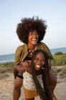 Cheerful young African American female with Afro pigtails giving piggyback ride to laughing girlfriend with curly hair while having fun during summer holidays together on seashore — Stock Photo