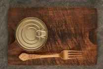 From above scratched chopping board with fork and sealed can with preserved food on rustic lumber table — Stock Photo