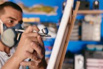 Vista lateral de artista masculino recortado en respirador usando pistola de pulverización para pintar el cuadro en lienzo durante el trabajo en taller creativo - foto de stock