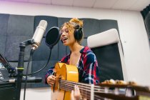 Молодая вокалистка в наушниках, играющая на акустической гитаре, поет с закрытыми глазами в микрофон в музыкальной студии — стоковое фото