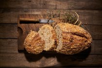 Вид на аппетитный свежеиспеченный домашний хлеб с хрустящей корочкой и овсяными хлопьями на деревянном столе с ножом и ароматическими травами — стоковое фото