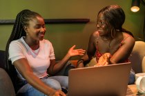 Alegres colegas afroamericanas navegando por la computadora portátil mientras trabajan de forma remota en la cafetería y discuten el proyecto empresarial - foto de stock