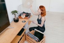 Junge Frau wählt Schallplatte aus Holzcontainer aus, während sie im Hauszimmer gegen Vintage-Plattenspieler sitzt — Stockfoto