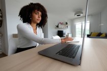 Vista lateral de la freelancer afroamericana sentada en la mesa con el portátil y escribiendo en el bloc de notas mientras trabaja remotamente en el proyecto en casa - foto de stock