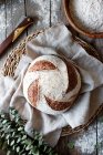 De arriba hecha en casa masa fermentada fresca pan de espelta en soporte de mimbre con tela sobre mesa de madera con harina dispersa - foto de stock