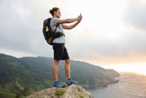 Side view of delighted male hiker taking self shot on smartphone while standing on hill on background of sea during trekking in summer — Stock Photo