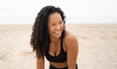 Позитивная азиатская спортсменка с вьющимися волосами, смеющаяся на песчаном побережье летом — стоковое фото