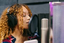 Conteúdo jovem vocalista feminina negra em fones de ouvido tocando barriga enquanto canta no microfone no estúdio de música — Fotografia de Stock
