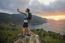 Vue latérale du randonneur masculin ravi de se prendre en photo sur smartphone tout en se tenant debout sur la colline sur fond de mer pendant la randonnée en été — Photo de stock