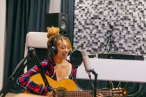Jovem vocalista étnica feminina em fones de ouvido tocando guitarra acústica enquanto cantava com os olhos fechados no microfone no estúdio de música — Fotografia de Stock