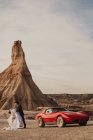 Bride tilting back in hands of groom while dancing near red sports car and mountain on cloudy day in Bardenas Reales Natural Park in Navarra, Spain — Stock Photo