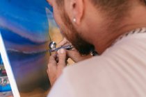 Side view of cropped unrecognizable male artist using spray gun to paint picture on canvas during work in creative workshop — Stock Photo