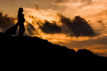 Baixo ângulo silhueta vista lateral de fêmea grávida anônima em pé sozinho na colina e sonhando com o futuro ao pôr do sol — Fotografia de Stock