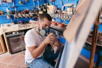 Vue latérale de l'artiste masculin dans un respirateur à l'aide d'un pistolet pulvérisateur pour peindre sur toile pendant le travail dans un atelier créatif — Photo de stock