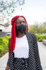 Jovem afro-americana em máscara protetora com dreadlocks brilhantes passeando no parque com mala e olhando para longe — Fotografia de Stock
