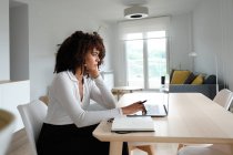 Vista laterale della freelance afroamericana seduta a tavola con computer portatile e scritta in blocco note mentre lavora a distanza sul progetto a casa — Foto stock