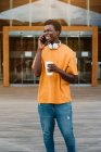 Улыбающийся афроамериканец с чашкой кофе счастливо смеется, разговаривая по мобильному телефону и отворачиваясь — стоковое фото
