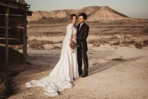 Full body glad groom in elegant suit embracing cheerful bride in white dress while standing near shed in Bardenas Reales Natural park in Navarra, Spain — Stock Photo