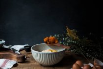 Tigela com ovos, farinha e colher de madeira com purê de abóbora na mesa de madeira durante a preparação da pastelaria no fundo escuro — Fotografia de Stock