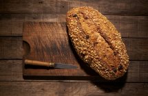 From above of appetizing crispy freshly baked homemade bread loaf with oatmeal flakes and raisin placed with knife on wooden cutting board — Stock Photo
