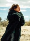 Vue latérale de la petite fille souriante enveloppée dans un manteau de fourrure avec les yeux fermés debout dans le vent sur une plage de sable fin par temps ensoleillé — Photo de stock