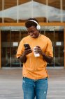 Fröhlicher afroamerikanischer Mann surft auf Smartphone und hört Musik über Kopfhörer, während er gegen ein modernes Gebäude läuft — Stockfoto