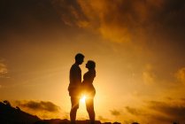 Low angle side view silhouettes of unrecognizable couple expecting baby standing looking at each other holding hands while admiring sundown in mountains together — Stock Photo