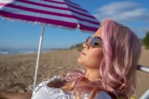 Вид збоку розслабленої жінки з рожевим волоссям, що лежить у кріслі та засмагає під час холоду на морі під час літніх канікул — стокове фото