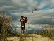 Vista laterale della madre viaggiatrice irriconoscibile con bambino sulle mani in piedi su un terreno collinare al mare contro il cielo nuvoloso con aquilone volante — Foto stock