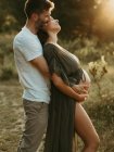 Seitenansicht eines lächelnden Mannes, der schwangere Frau von hinten umarmt, während sie bei Sonnenuntergang auf der Wiese steht — Stockfoto