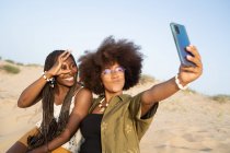 Giovani amiche afroamericane allegre che sorridono brillantemente mentre scattano selfie sullo smartphone durante il fine settimana estivo sulla spiaggia sabbiosa — Foto stock