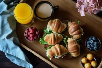 Вкусные бутерброды с круассаном и овощами, подаваемые на подносе с капучино и апельсиновым соком, приготовленные на французский завтрак и помещенные на деревянный стол — стоковое фото