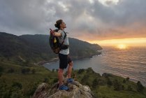 Seitenansicht eines unbeschwerten männlichen Wanderers, der am Aussichtspunkt auf einem Felsen steht und die Freiheit vor dem Hintergrund des Sonnenuntergangs über dem Meer genießt — Stockfoto