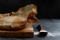 Состав из свежеиспеченного рустикального теста круглого хлеба на пергаментной бумаге, помещенный на деревянную доску с ложкой и пшеничной мукой — стоковое фото