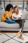 Ganzkörperlachendes junges lesbisches Paar blättert gemeinsam im Netbook, während es auf dem gemütlichen Sofa im Wohnzimmer sitzt — Stockfoto