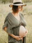 Спокойная беременная женщина в платье и соломенной шляпе трогает животик, стоя на поле в сельской местности на закате летом, глядя в сторону — стоковое фото