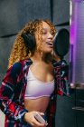 Contenida joven vocalista negra en auriculares tocando el vientre mientras canta en el micrófono en el estudio de música - foto de stock