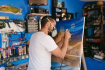 Mann unter Atemschutz sprüht Farbe auf Leinwand mit abstrakter Landschaft bei der Arbeit im professionellen Kreativatelier — Stockfoto