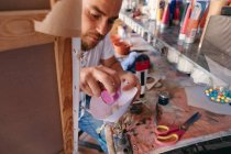 Alto ángulo de artista masculino vertiendo pigmento líquido en pistola de pulverización mientras está sentado cerca de la mesa detrás del caballete con lienzo en el taller - foto de stock