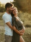 Vue latérale d'un homme souriant embrassant une femelle enceinte par derrière alors qu'il se tenait debout dans un pré à la campagne au coucher du soleil — Photo de stock