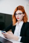Junge Unternehmerin in formeller Kleidung trägt SMS auf dem Handy, während sie zu Hause am Schreibtisch mit Laptop arbeitet — Stockfoto