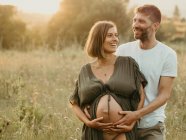 Sonriente macho abrazando a la hembra embarazada por detrás mientras está de pie en el prado del campo al atardecer mirando hacia otro lado - foto de stock
