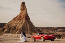 Невеста наклоняется назад в руках жениха во время танцев возле красного спортивного автомобиля и горы в пасмурный день в природном парке Барденас-Реалес в Наварре, Испания — стоковое фото