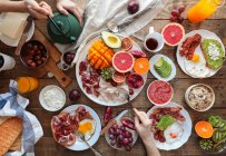 From above wide view of wooden table with multiple healthy meals prepared for breakfast. Crop hands with kitchenware — Stock Photo