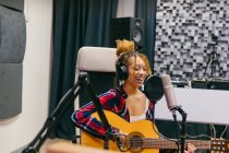 Giovane cantante etnica femminile in cuffia che suona la chitarra acustica mentre canta a occhi chiusi nel microfono in studio musicale — Foto stock