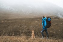 Side view of hiking woman in bright blue jacket and backpack with brown dog in dry empty valley surrounded by snowy mountains — Stock Photo